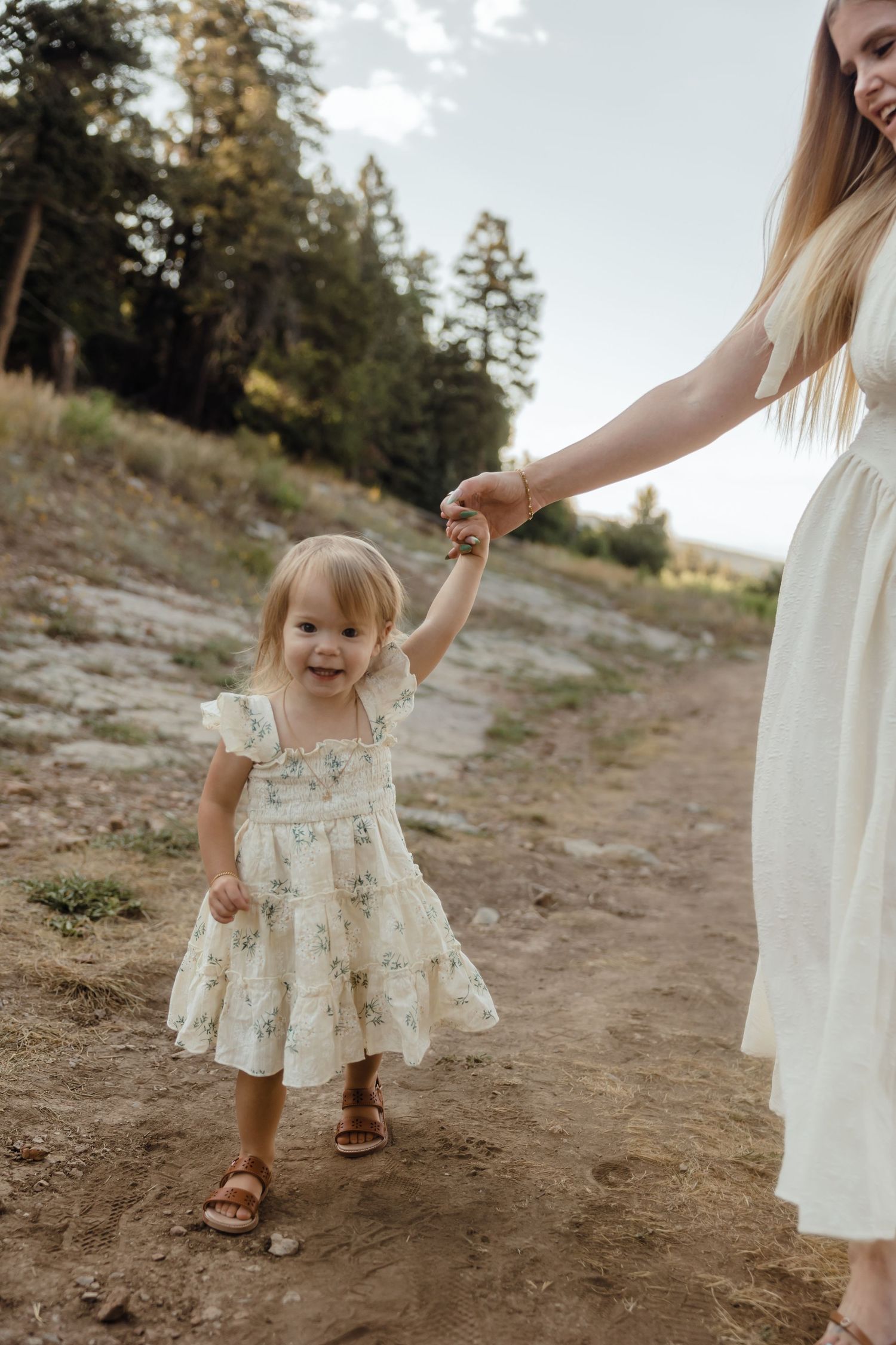 A toddler girl in a floral dress holds her mother's hand on a dirt path surrounded by pine trees.