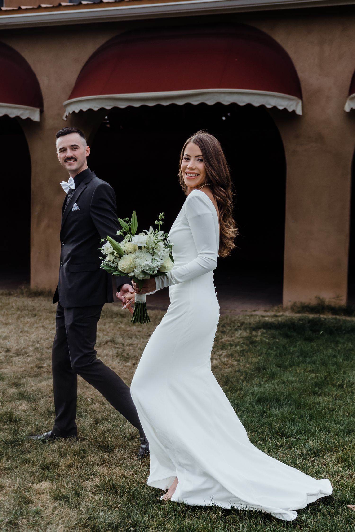 Bride in elegant white gown holds bouquet while walking with groom in black tuxedo outdoors.