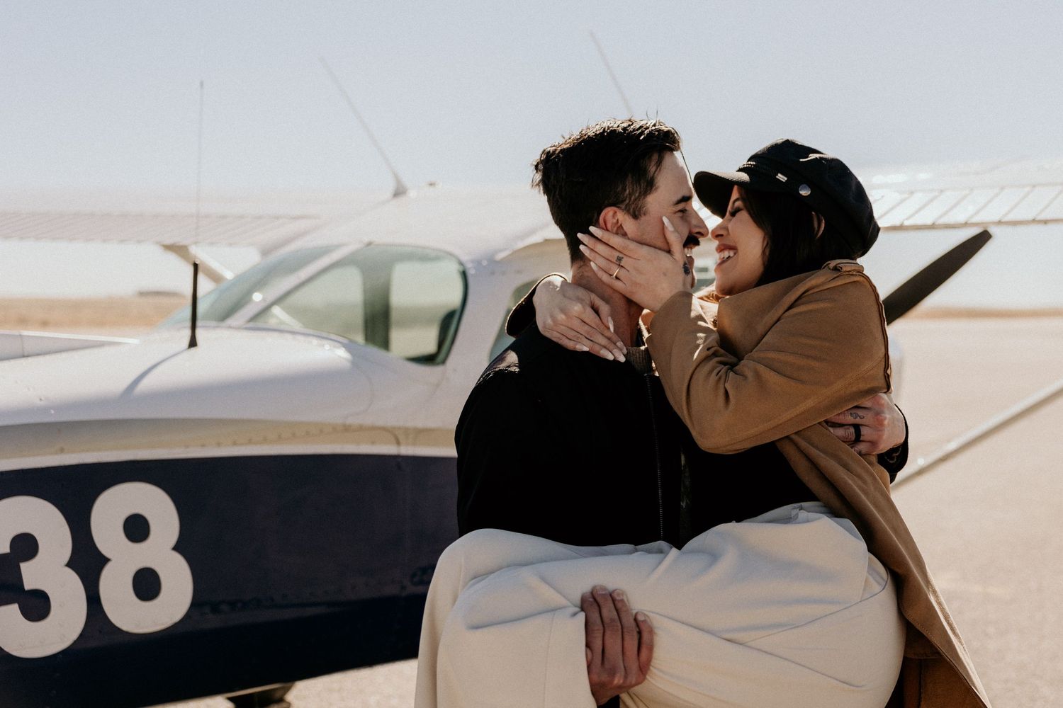 A couple shares a romantic kiss beside a small propeller plane on a sunny airstrip.