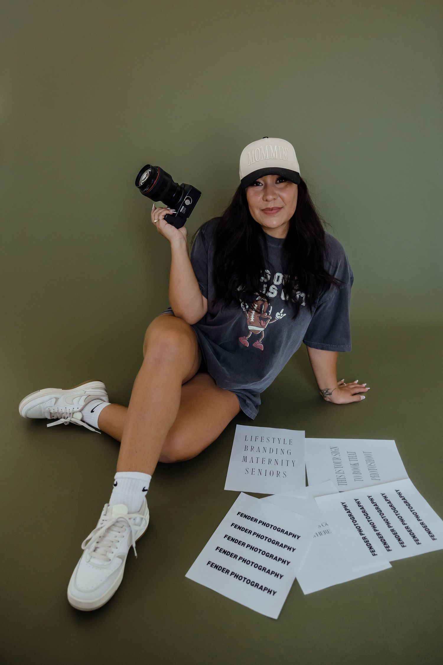 A female photographer sits against an olive green wall holding a camera surrounded by Fender Photography branding sheets.