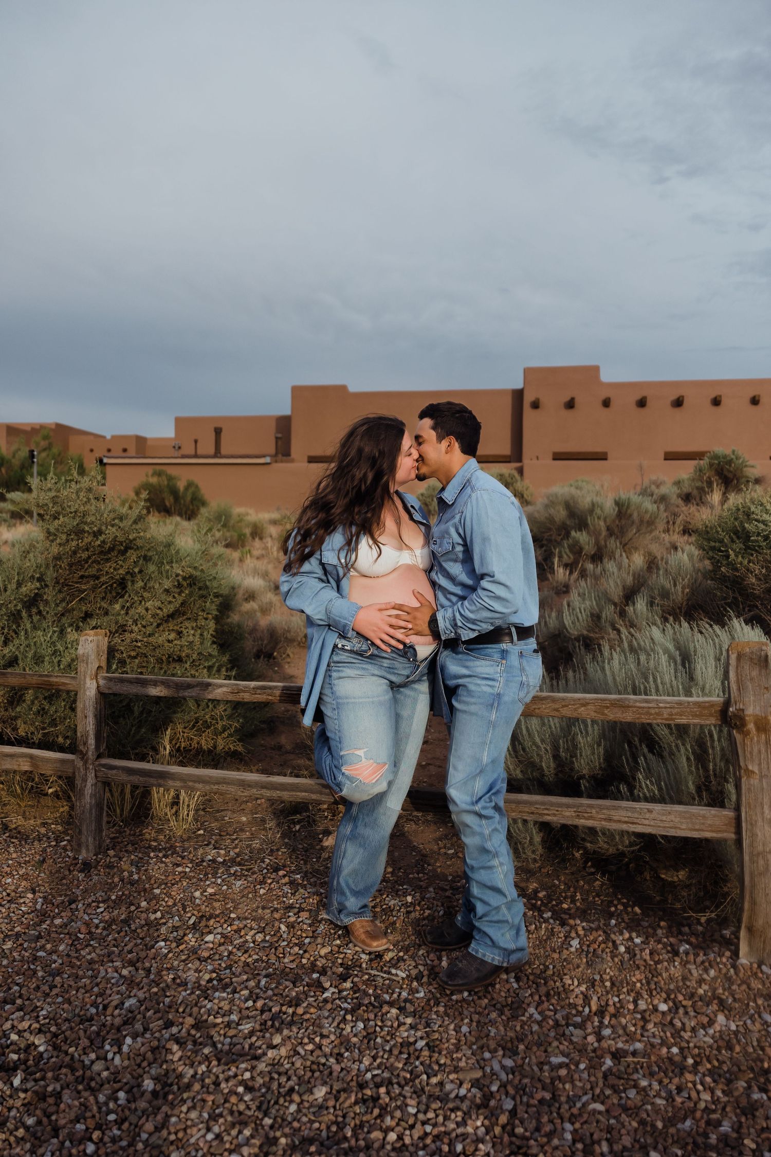 Expecting couple in matching denim kissing near a wooden fence with adobe building in background.