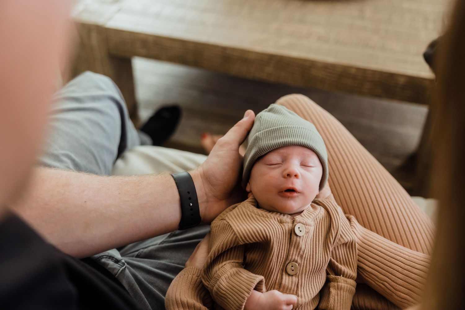 Newborn baby sleeping peacefully in a tan ribbed outfit and sage green hat, cradled in an adult's arms.