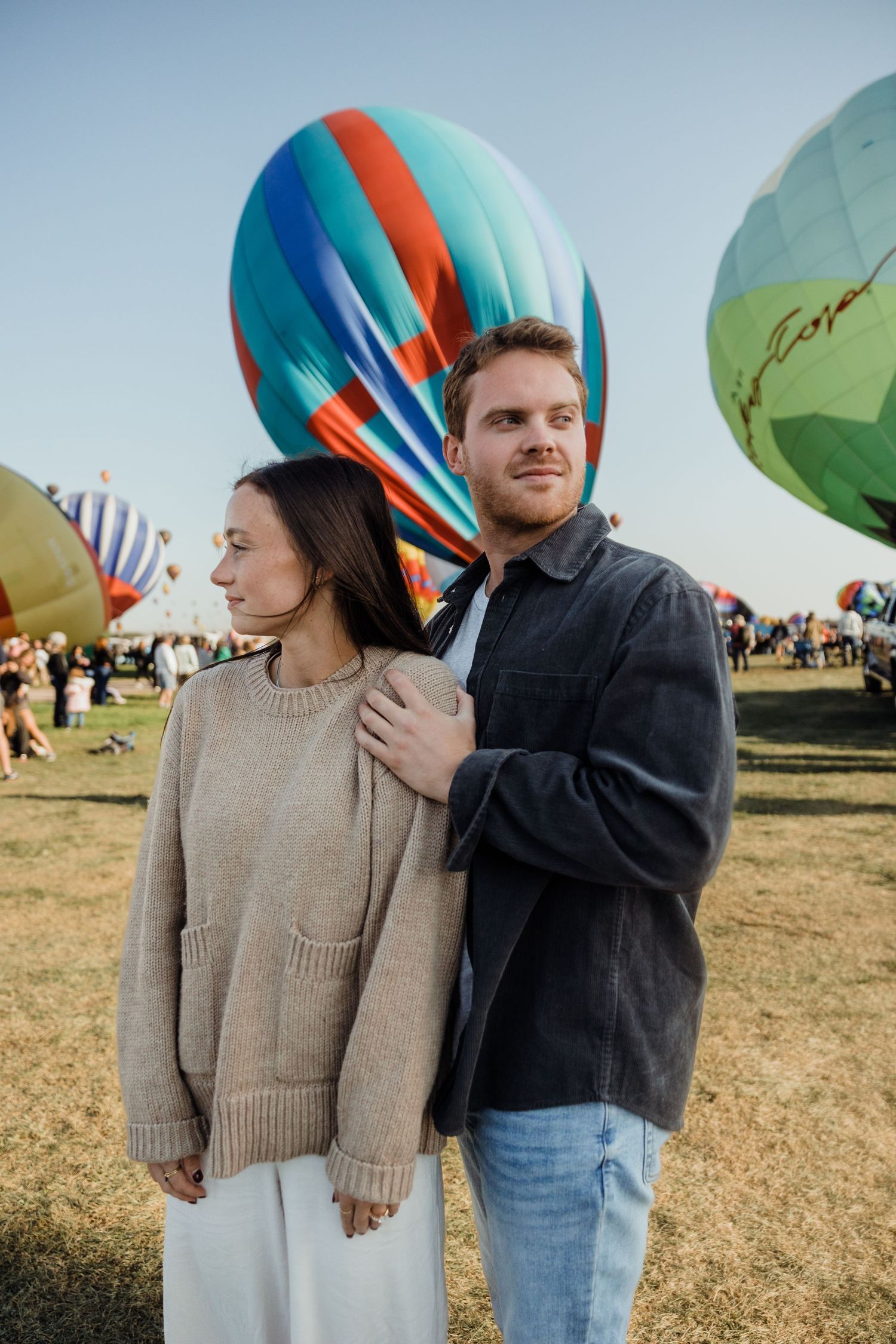 A couple stands together at a colorful hot air balloon festival on a sunny day.