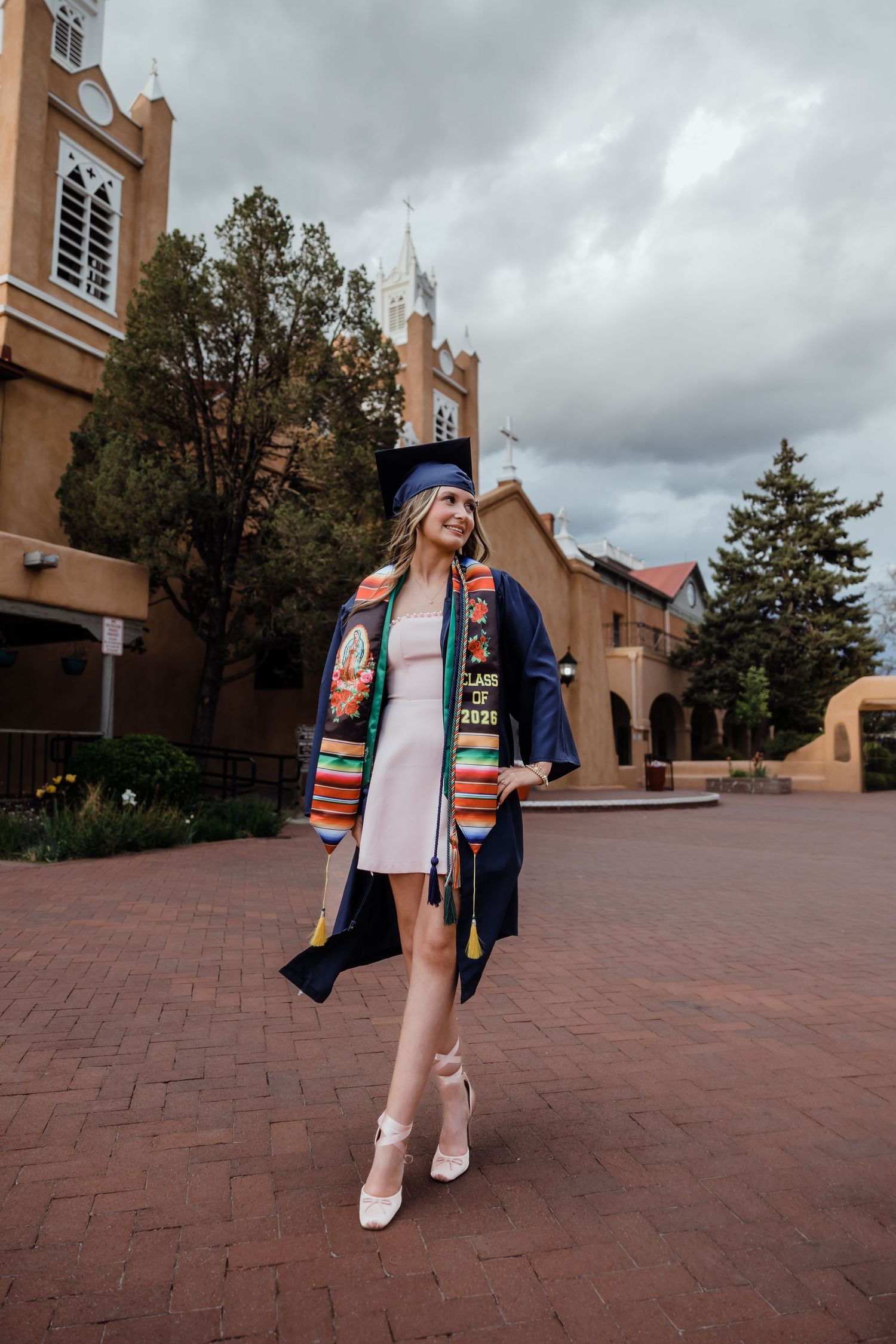 A smiling graduate poses in cap, gown, and colorful stole outside a historic adobe building on a cloudy day.
