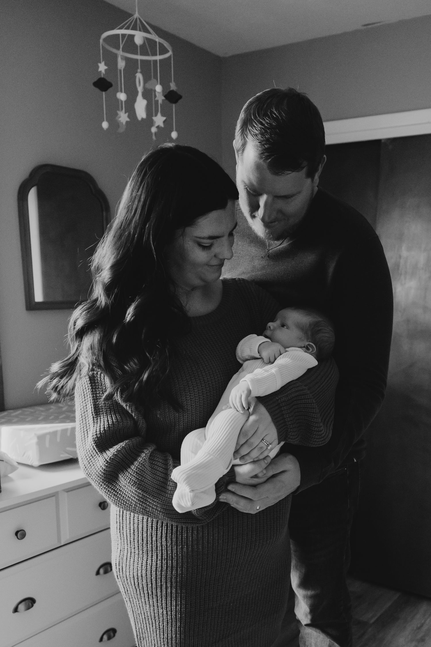 Black and white photo of new parents lovingly gazing at their newborn baby in a cozy nursery.