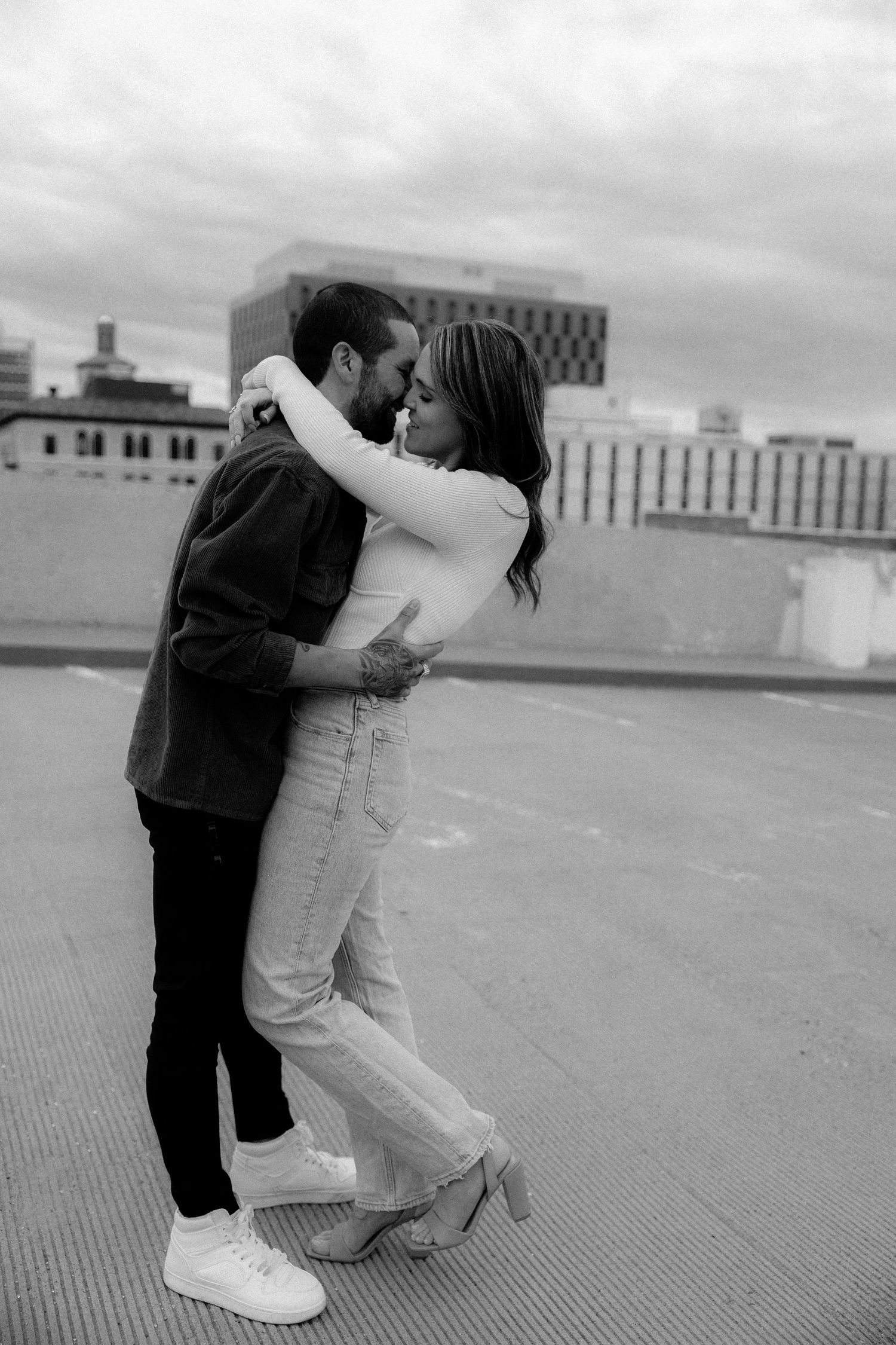 Black and white photo of a couple embracing and kissing on a rooftop with city buildings in background.