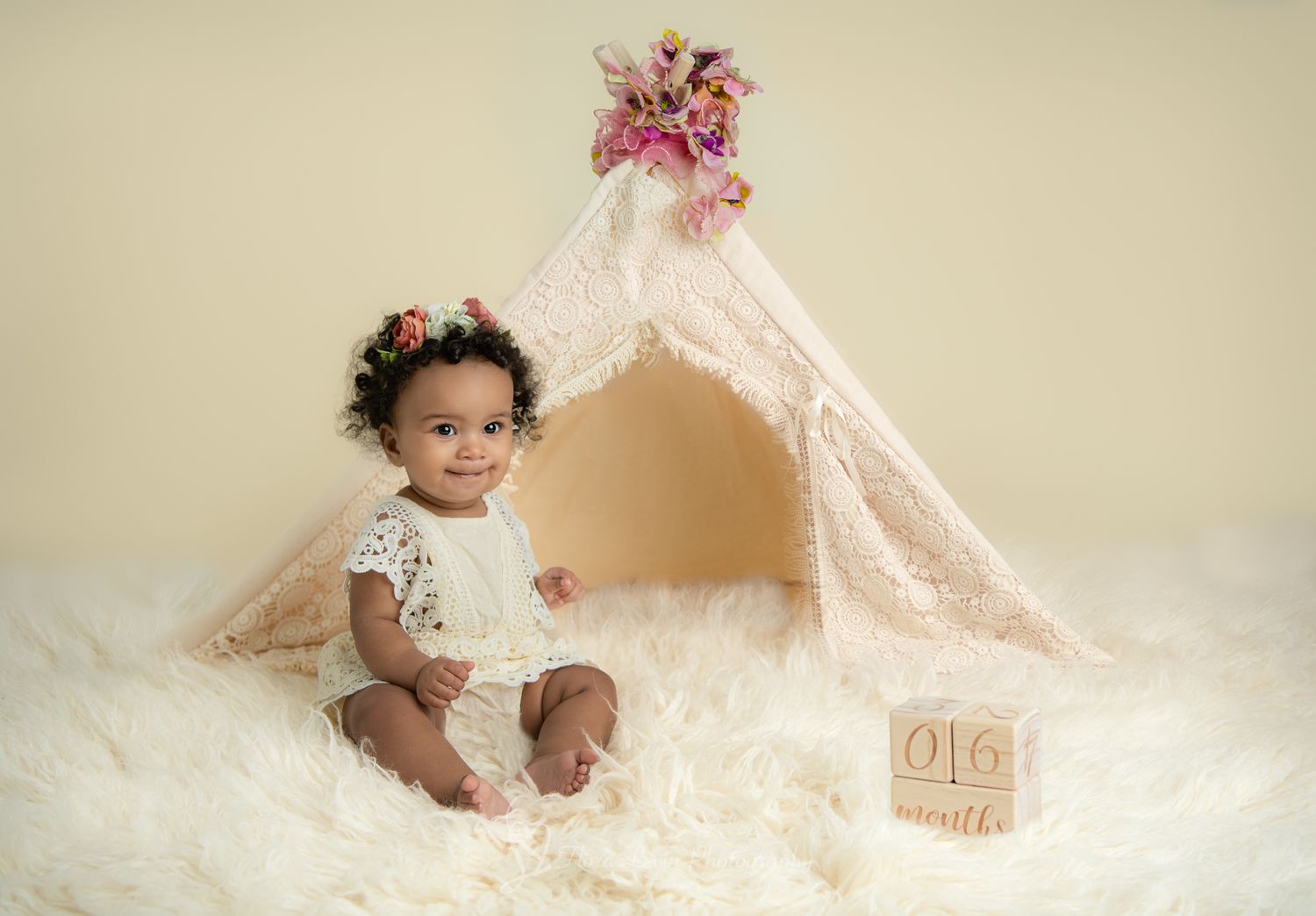 Smiling baby girl in white lace outfit sits on fluffy rug beside a decorative teepee with floral accents and milestone blocks.