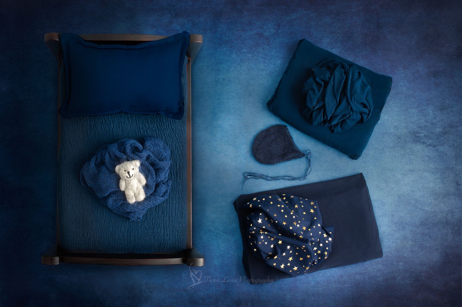 Navy blue newborn photography props flatlay with tiny bed, teddy bear, knit wraps, star blanket, and heart hat.