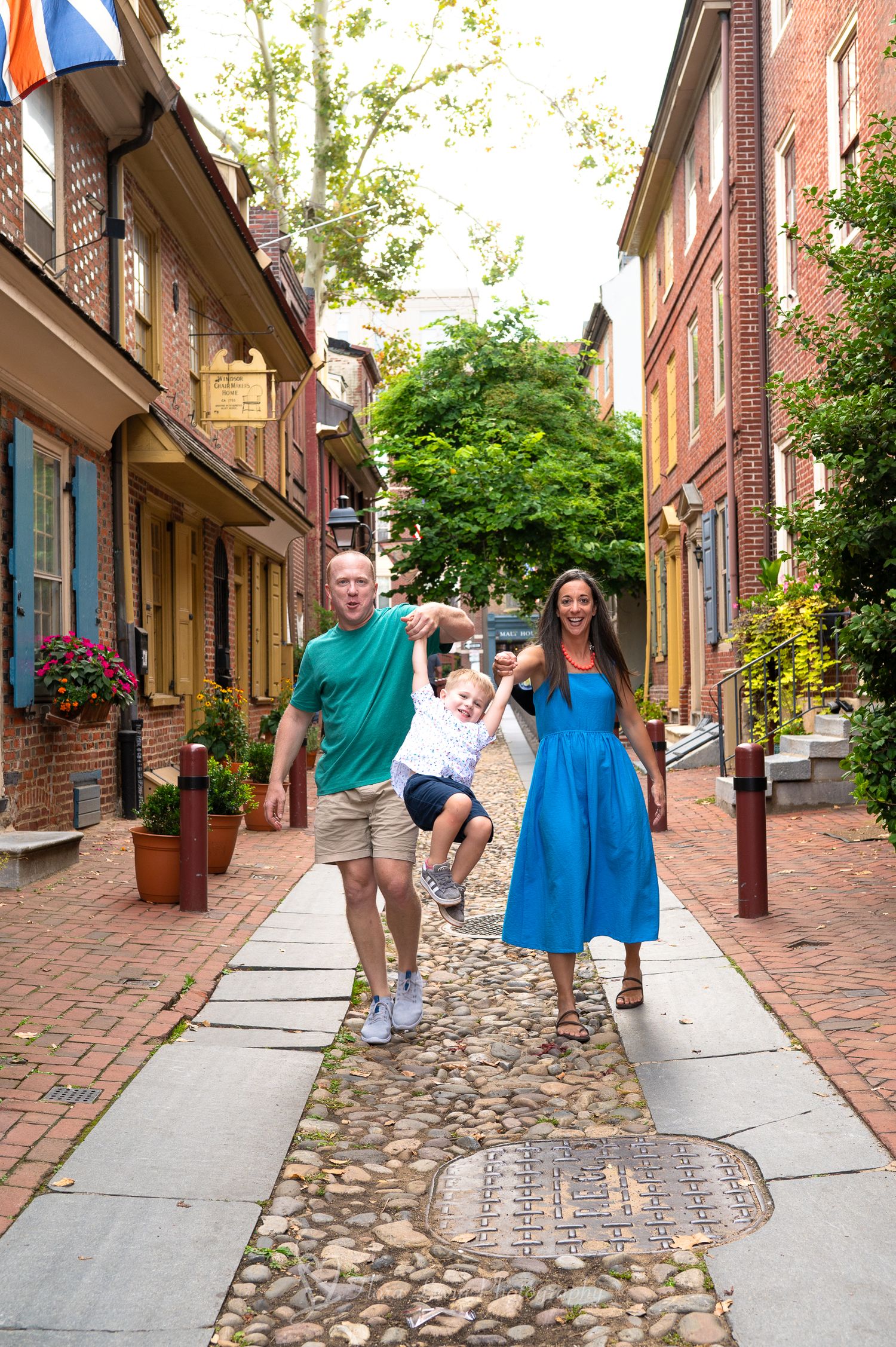 A family walks playfully along a Philadelphia historic cobblestone street lined with brick buildings and flower boxes.