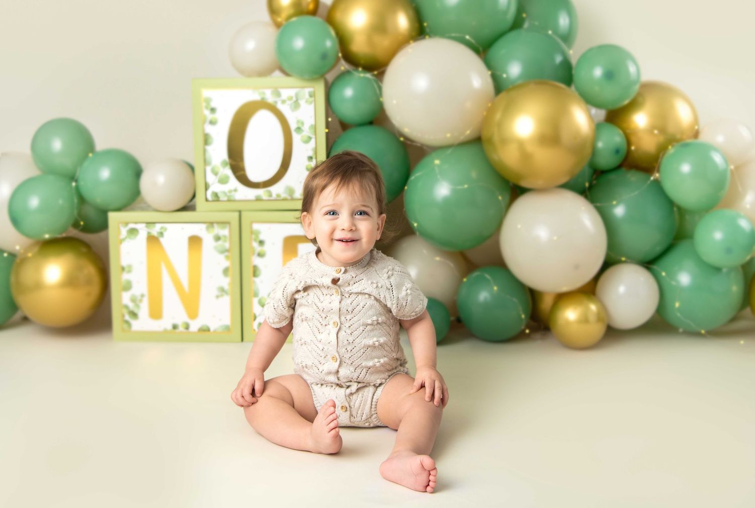 A first birthday setup with mint green, gold and white balloon garland against a cream colored backdrop. Baby boy wearing a neutral outfit.