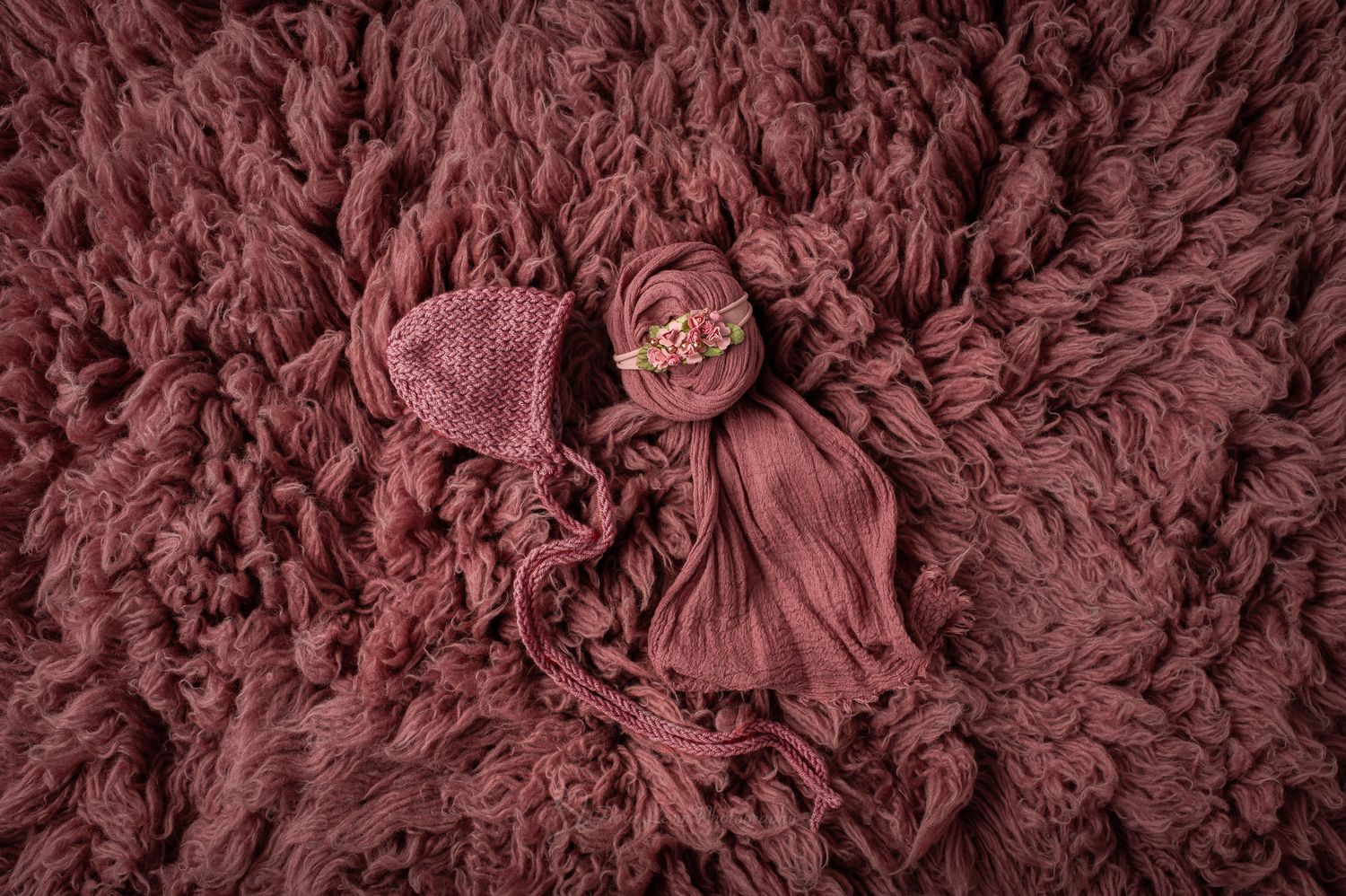 A newborn baby wrapped in a dusty rose knit bonnet and wrap, nestled on a matching textured flokati rug.