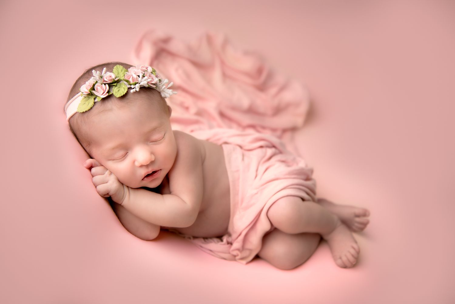 A sleeping newborn baby girl  lies peacefully in soft pink blanket, covered with a soft pink blanket, wearing delicate floral headband.