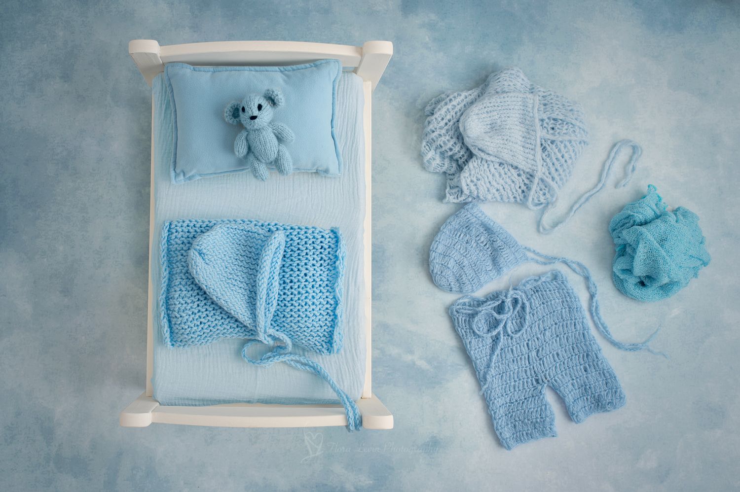 Blue knitted newborn baby clothes and accessories arranged with a tiny teddy bear on a  white wooden bed.