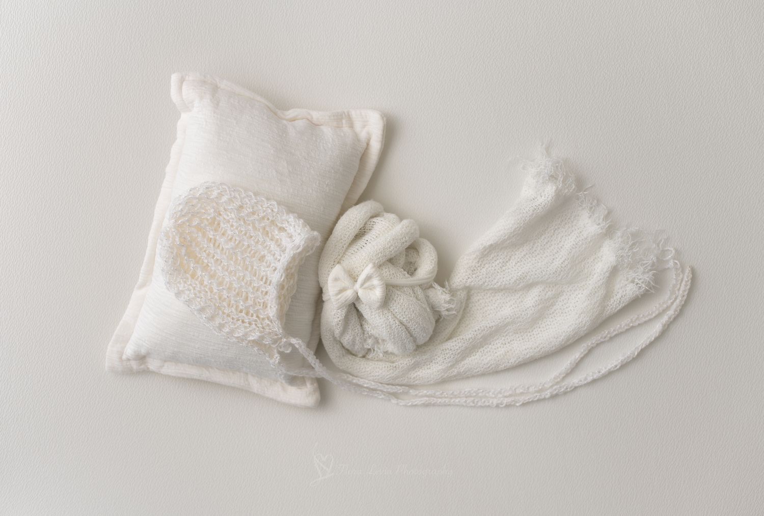 White newborn photography props including a knit bonnet, tiny booties, a pillow, and soft wrap on a white background.