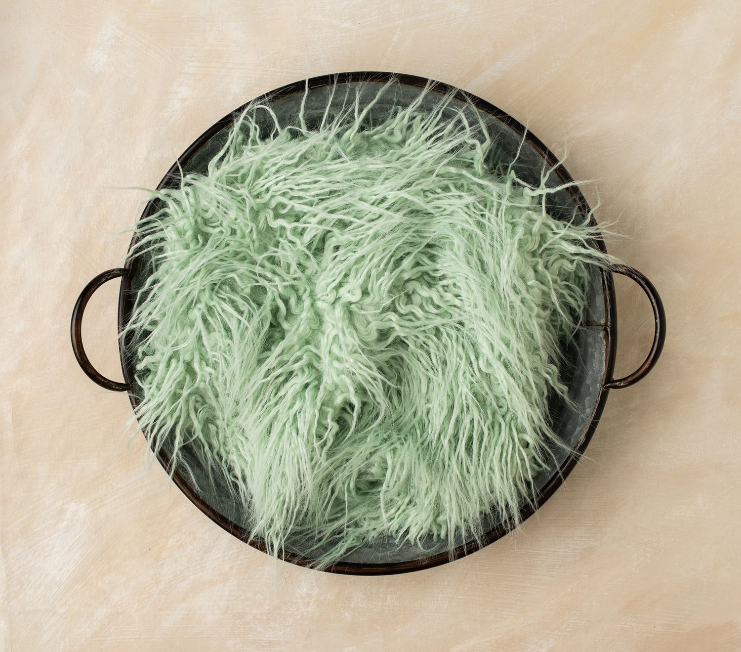 Soft mint green fluffy faux fur fabric draped in a dark round metal tray with handles on a beige background.