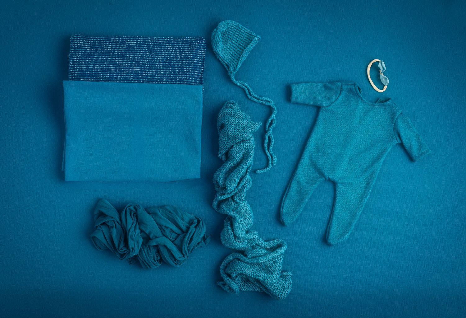 Teal newborn photography props set including a knit hat, headband, wrap, onesie, and fabric layers on a blue background.