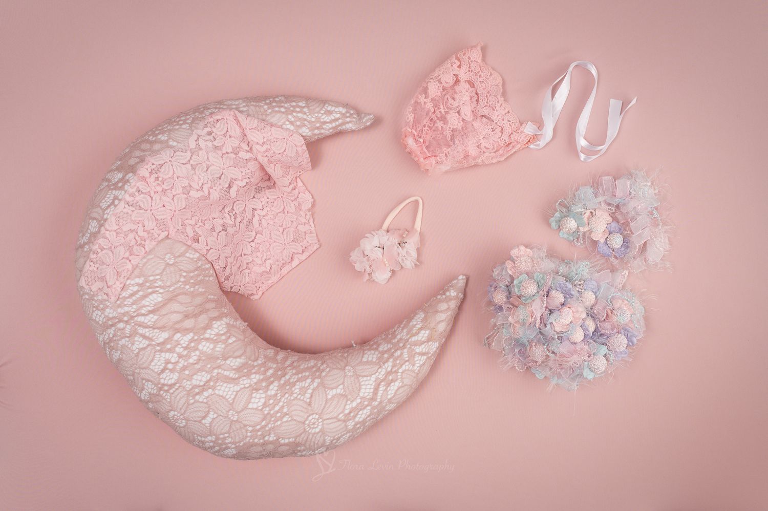 Pink newborn photography props including a crescent moon pillow, lace headband, bow clip, and floral bonnet on pink background.