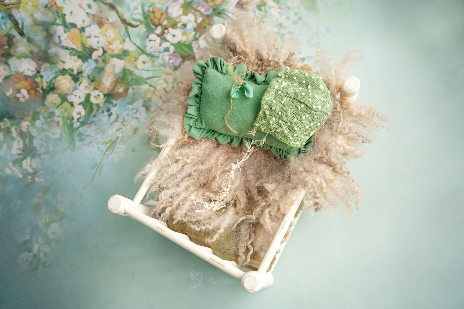 Newborn photography prop setup with green lace bonnet and outfit on fluffy beige nest in white mini bed.