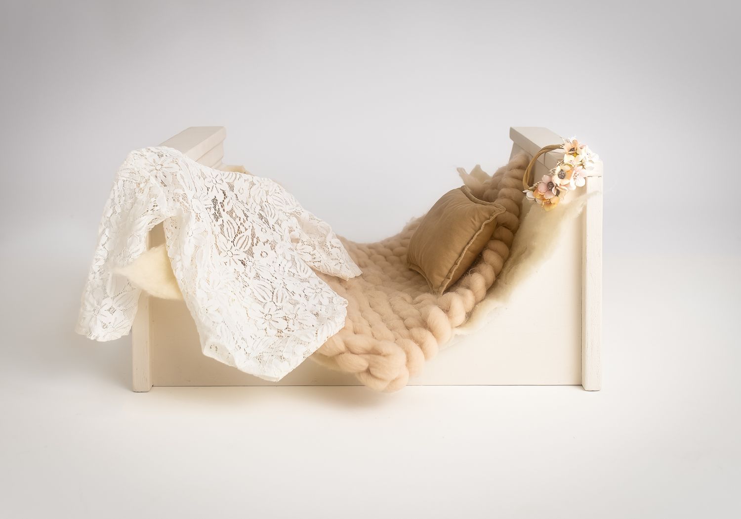 white wooden newborn arch bed prop with cream knit blanket, tan pillow and delicate lace romper and floral headband.