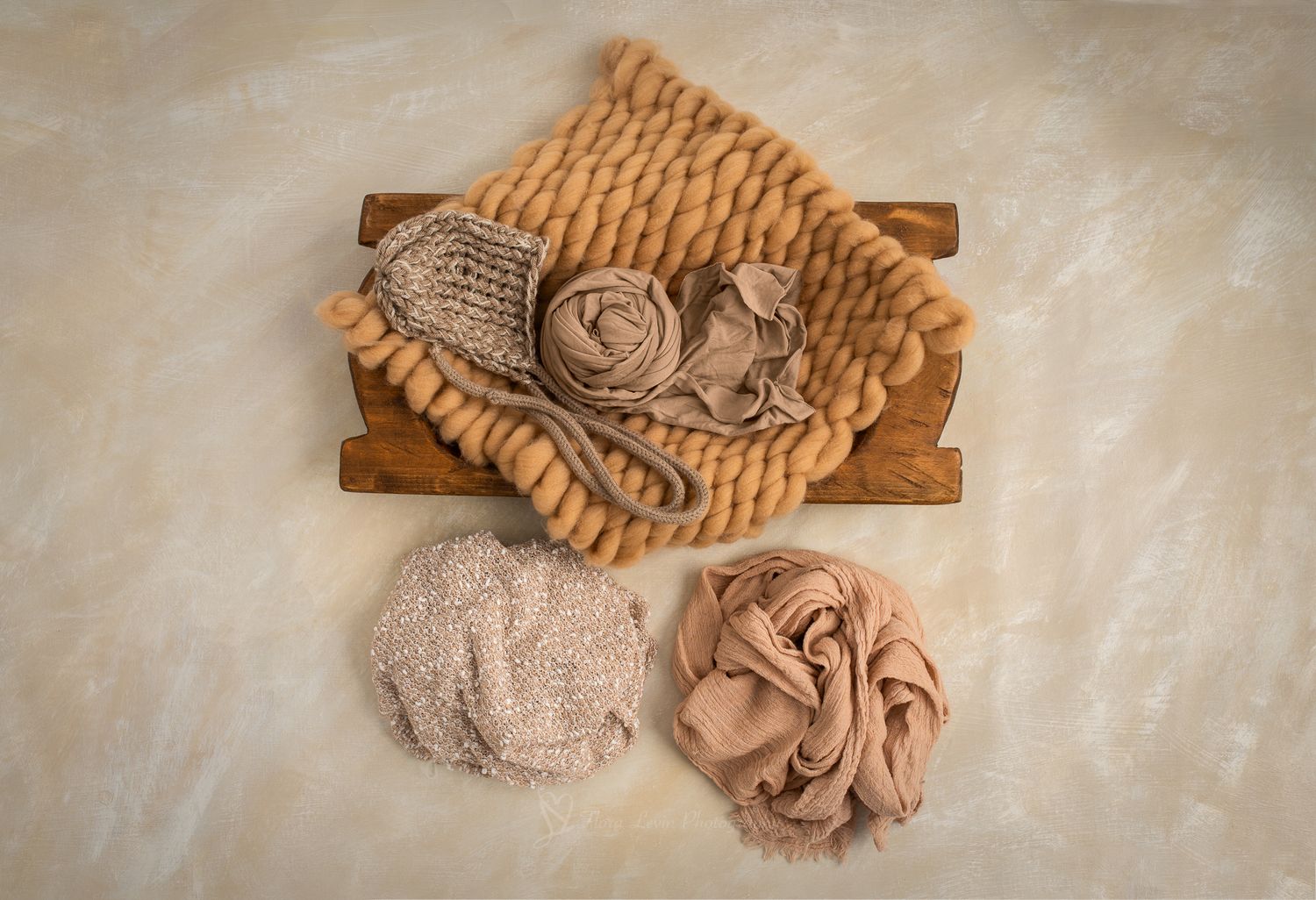 Newborn photography props including a wooden tray, chunky knit blanket, tiny hat, and two fabric wraps in neutral tones.