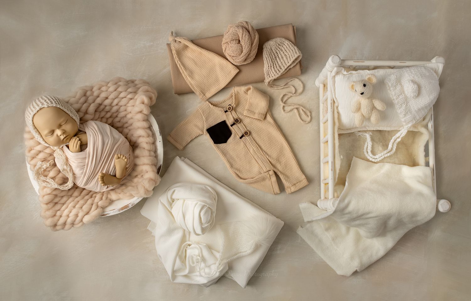 Newborn baby doll with cream knit props, beige outfit set, white blanket, and teddy bear  on neutral background.