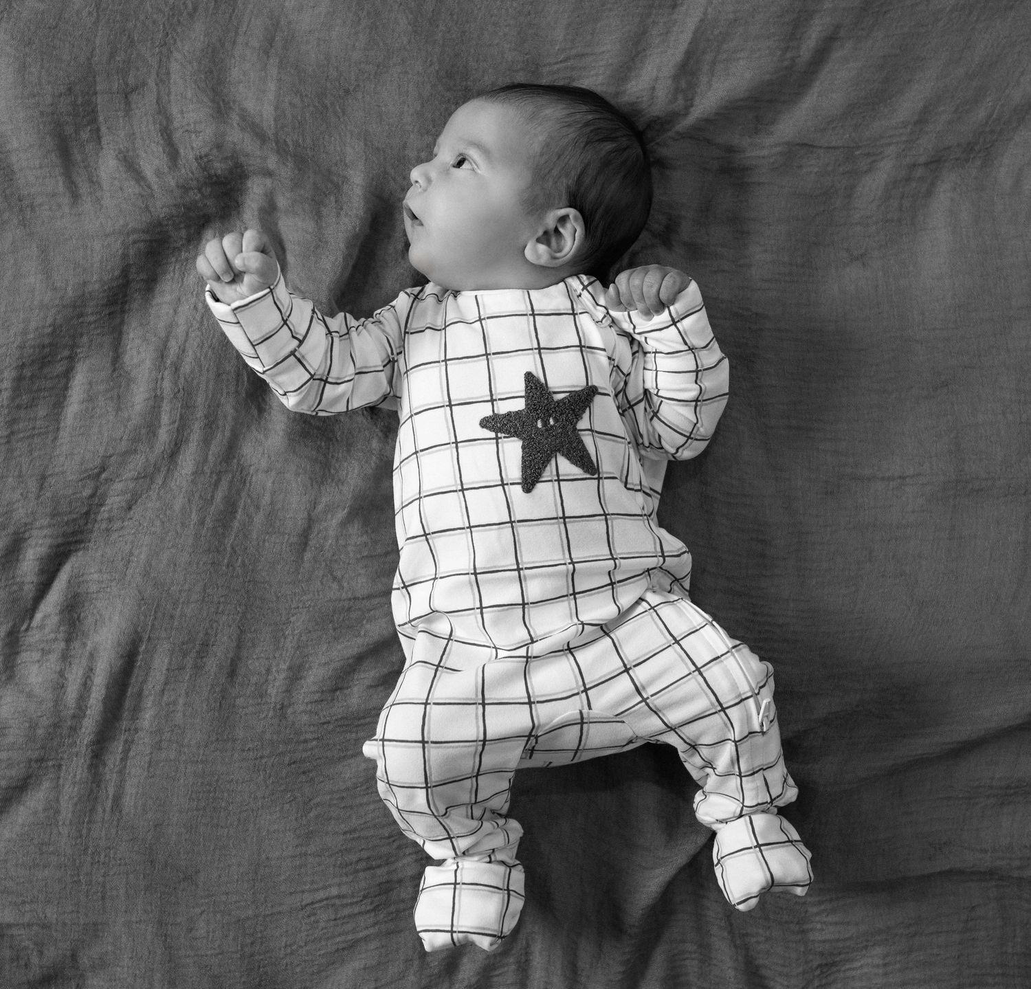 Baby in white and black checkered onesie lying on grey bedding shown in movement positions looking for his mom.