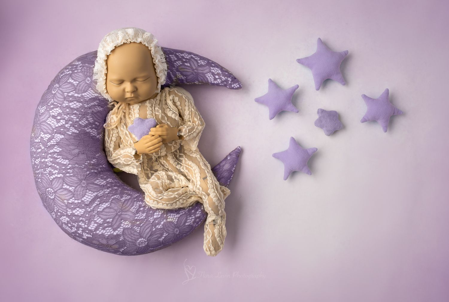 Baby doll in cream lace outfit resting on purple crescent moon prop with star decorations on lavender background.