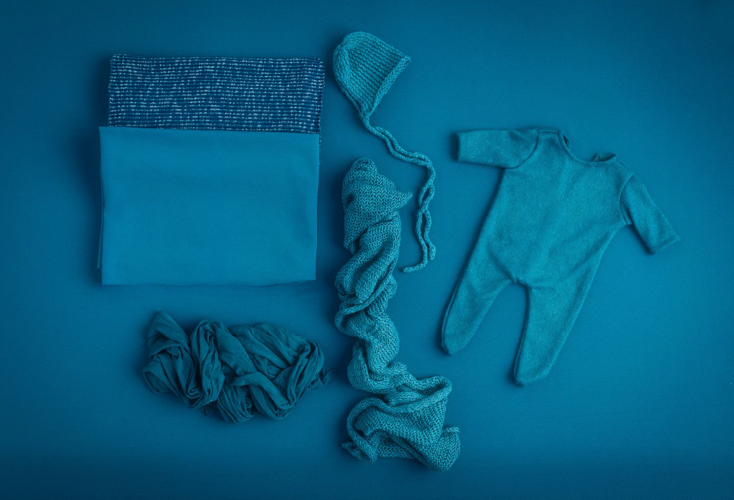 Teal baby clothing set with onesie, hat, wraps  arranged on blue background.