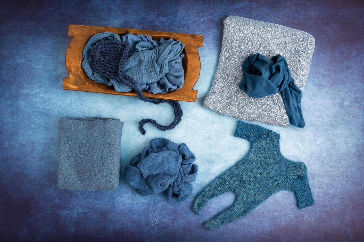 Flat lay of blue-gray knit newborn photography props including wraps, a onesie, and hat on a dark blue background.