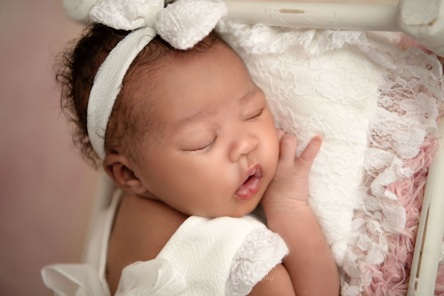 Sleeping newborn baby girl wearing a white bow headband and lace dress, peacefully resting on a soft pink  blanket.