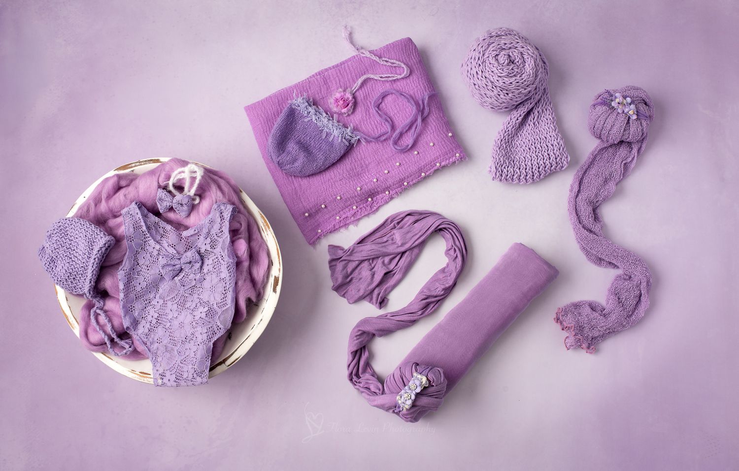 Purple newborn photography props including lace outfit, bonnet, wraps, and blanket on lavender background.