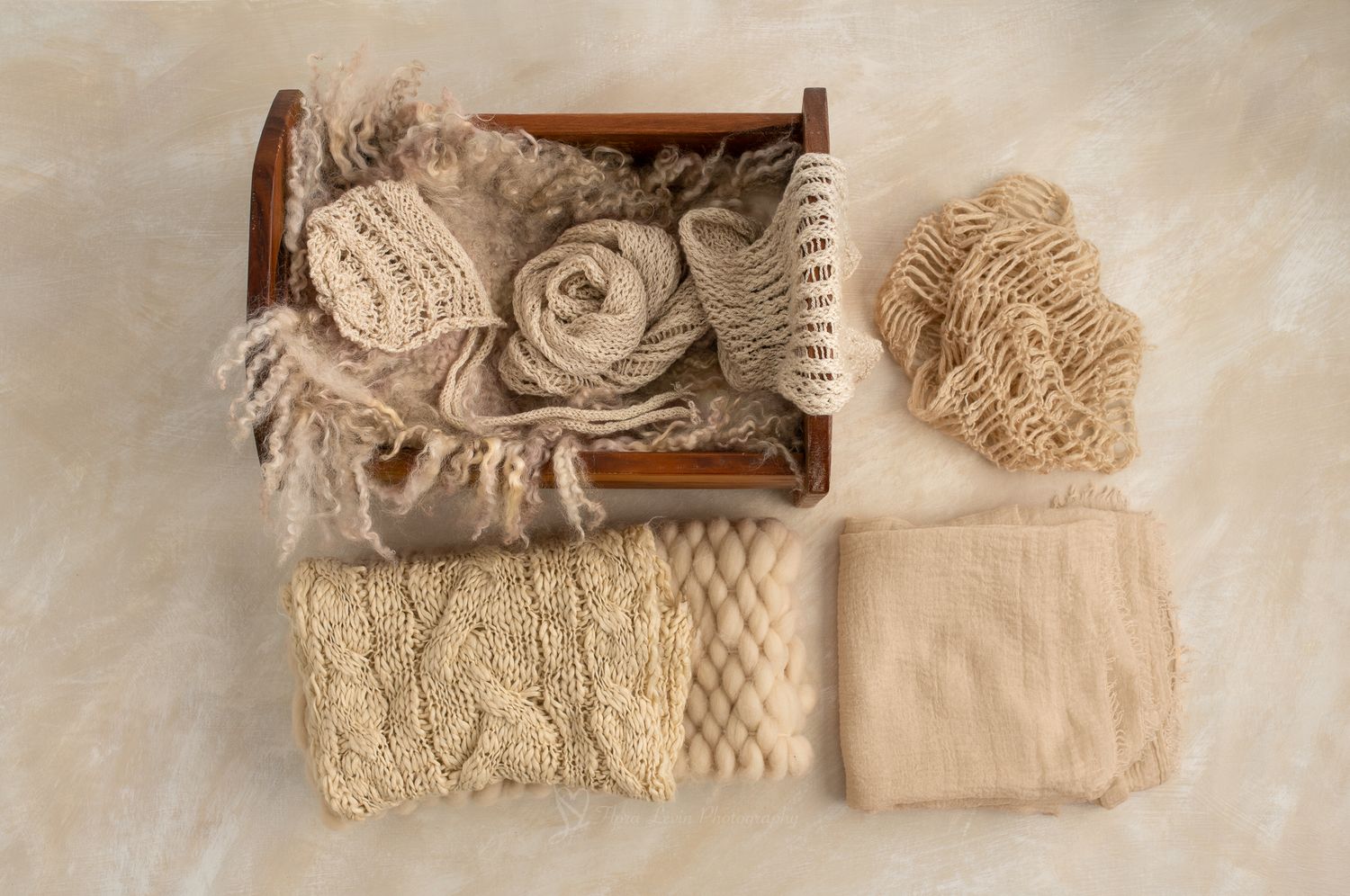 Neutral beige newborn photography props including knit wraps, lace layers, and a wooden  bed on a cream background.