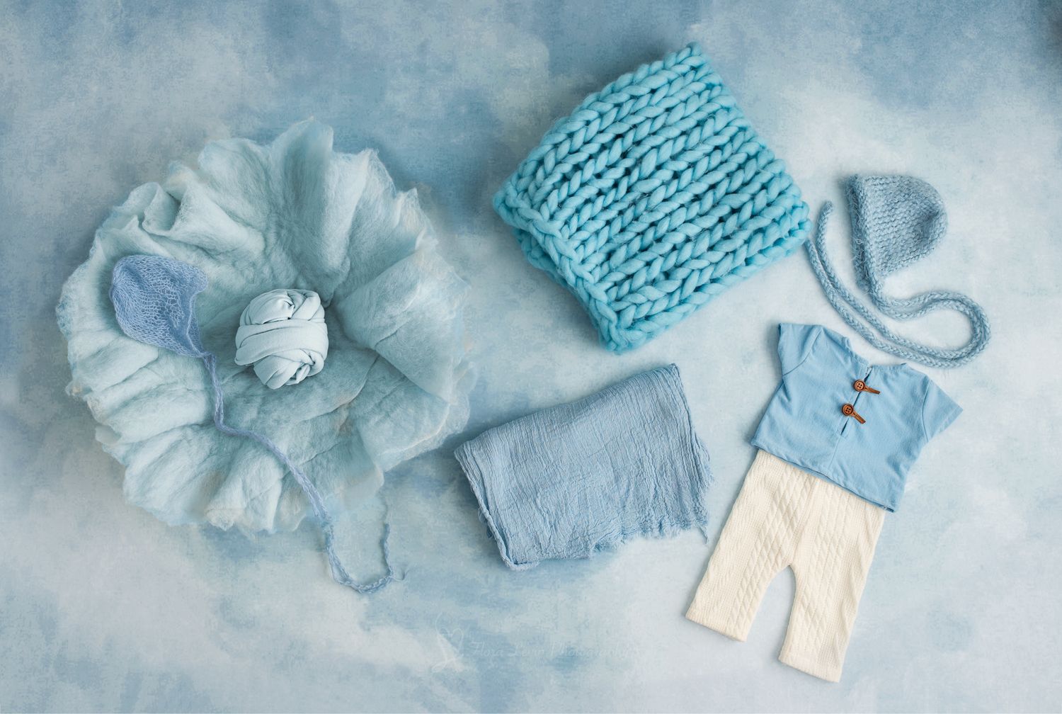 Blue newborn photography props including knit blanket, bonnet, outfit, and fabric layers on a soft blue background.