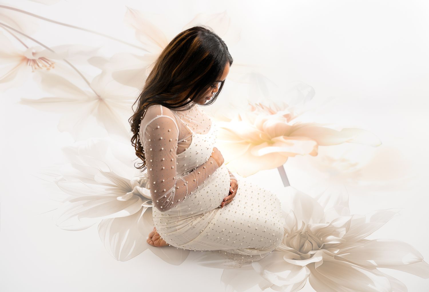 Pregnant woman in white lace dress sits gracefully among large floral decorations in a soft, dreamy maternity photo.