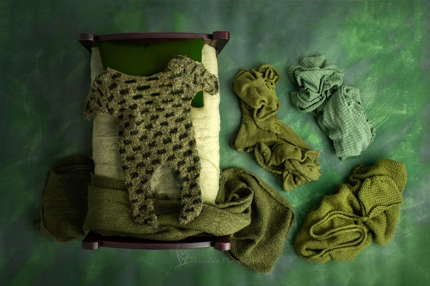Green knit newborn outfit on a miniature wooden bed surrounded by green fabric wraps on a green textured background.