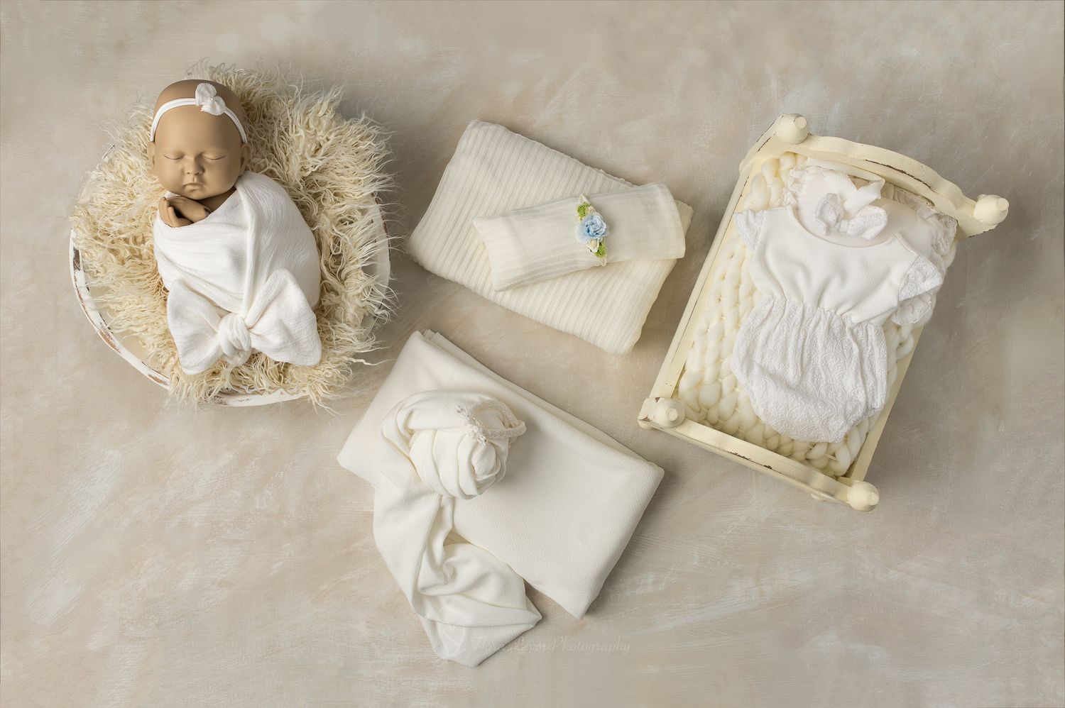 Newborn baby doll  swaddled in white wrap lying in wicker bowl beside folded ivory blankets and a miniature wooden crib.