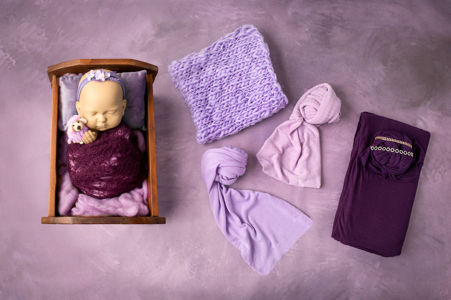 Newborn baby wrapped in dark purple lace in wooden bed beside lavender knit and purple fabric wraps.