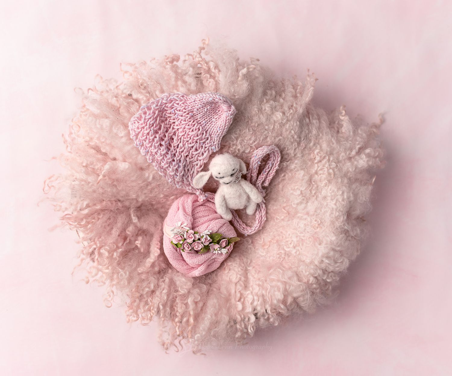 Newborn baby sleeping on pink fluffy blanket with knit hat and stuffed lamb toy in soft pink nursery setting.