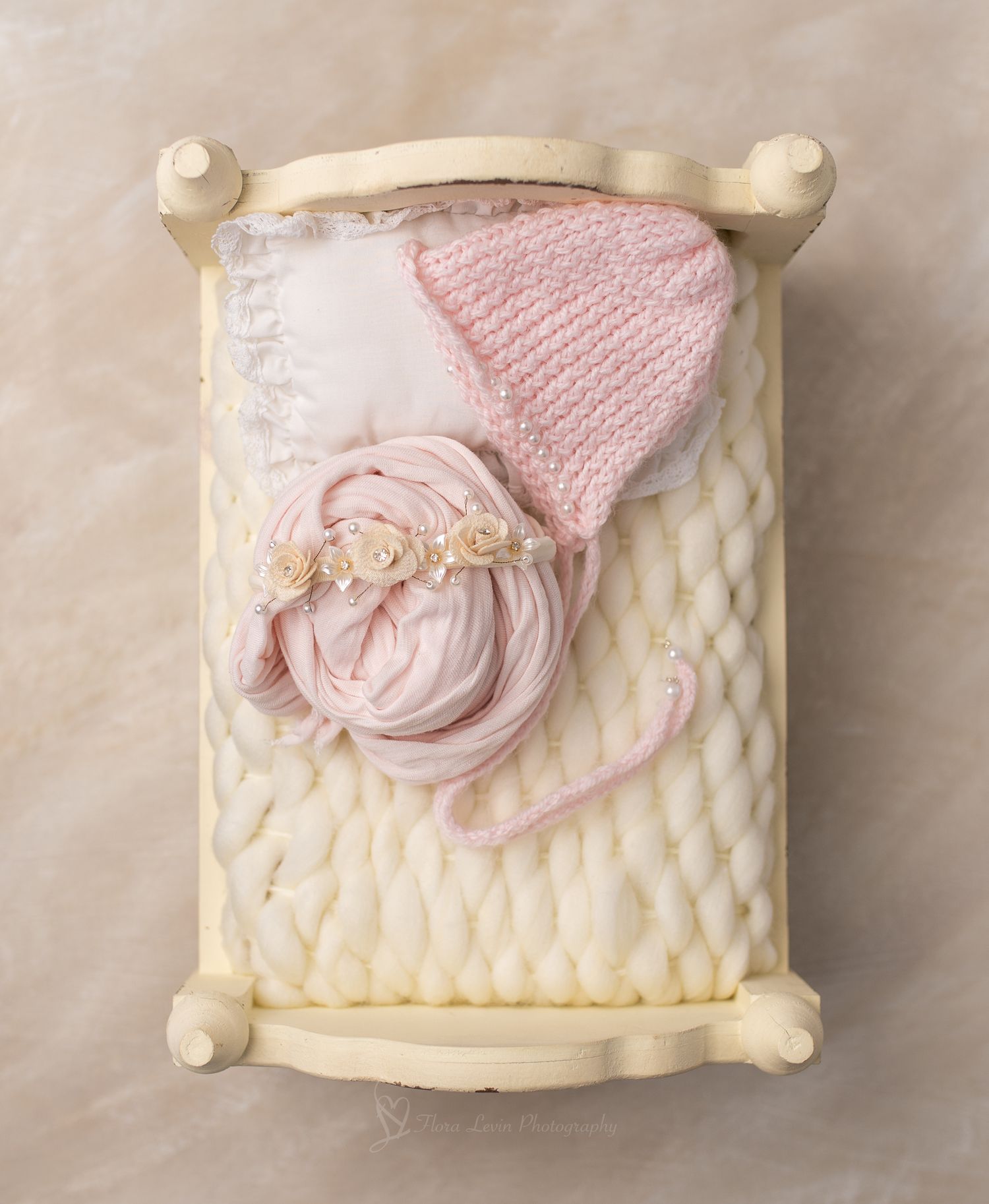 Pink knit newborn bonnet and floral headband on a miniature cream wooden bed with chunky knit blanket.