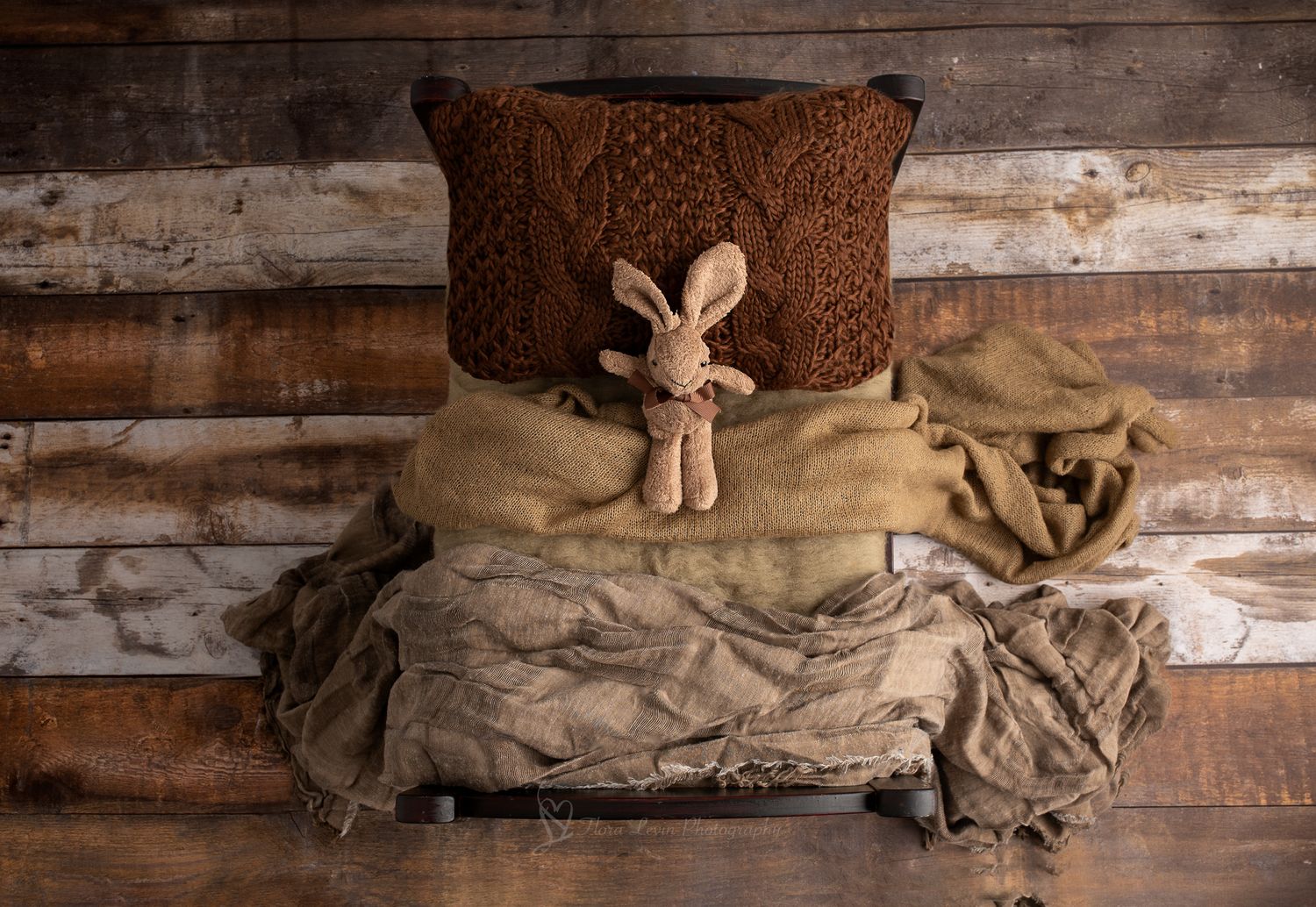 Rustic wooden backdrop with brown knit pillow, burlap blankets, and a small stuffed bunny toy on a vintage crib.
