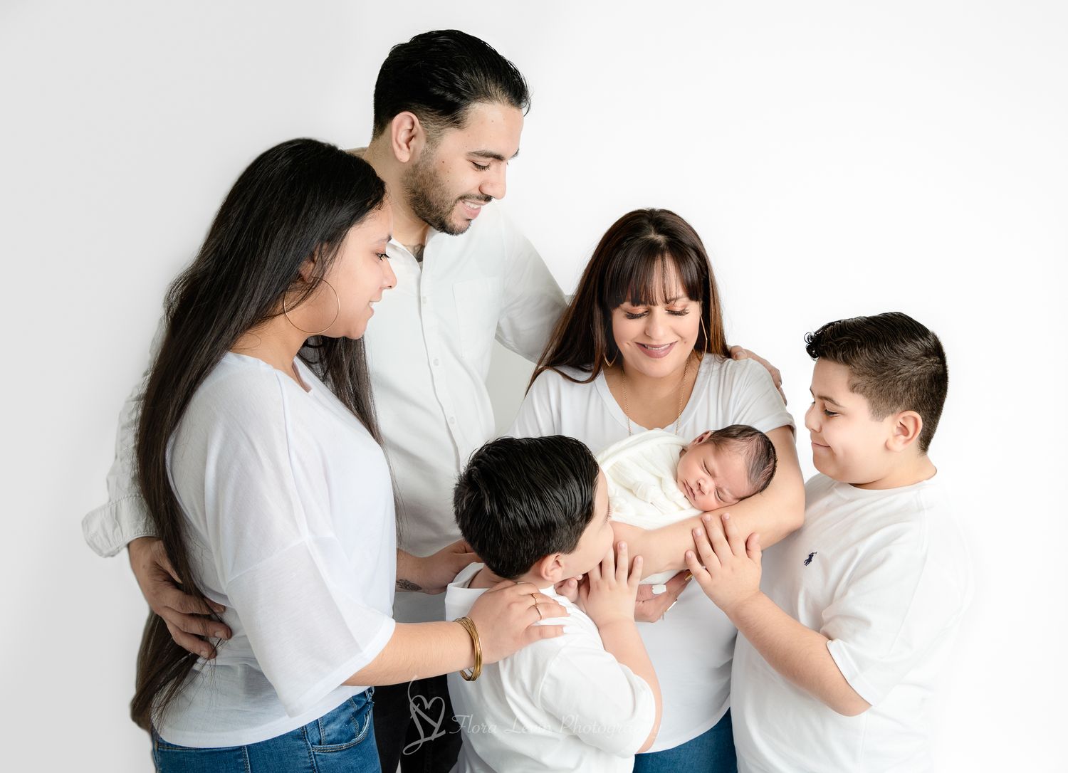 Family of six in white tops admiring their newborn baby against a clean white studio background.