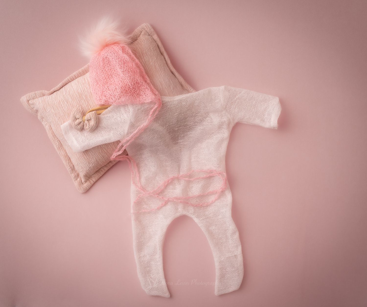 Pink newborn photography props including a fuzzy bonnet, tiny romper, and small pillow on a pink background.