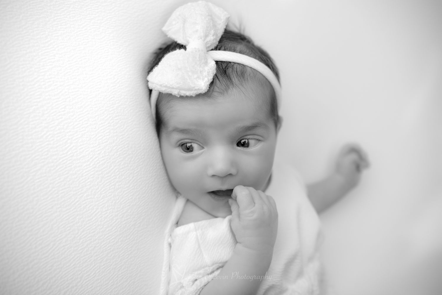 A baby wearing a white headband poses in four different black and white portraits.with her hands in her mouth.