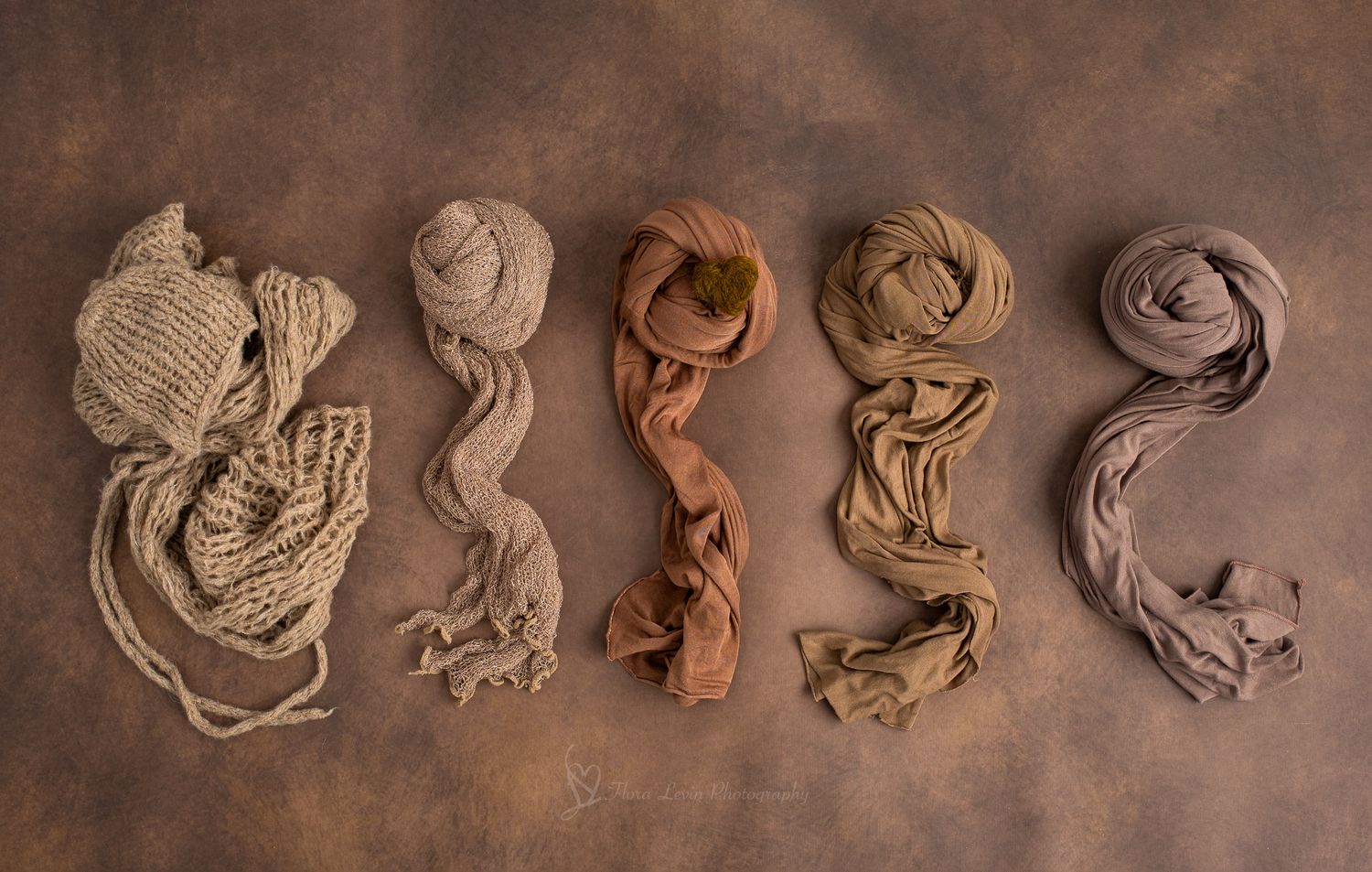 Five neutral-toned fabric wraps and yarn skeins arranged in a row on a brown textured background.