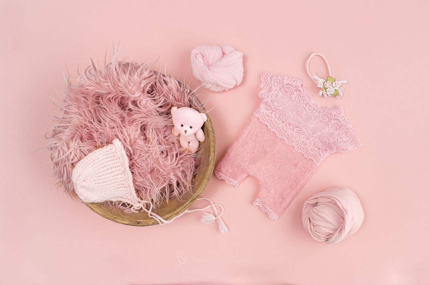 Pink newborn photography props including fluffy  fur in bowl, lace romper, knit hat, tiny bear, and wrap on pink background.