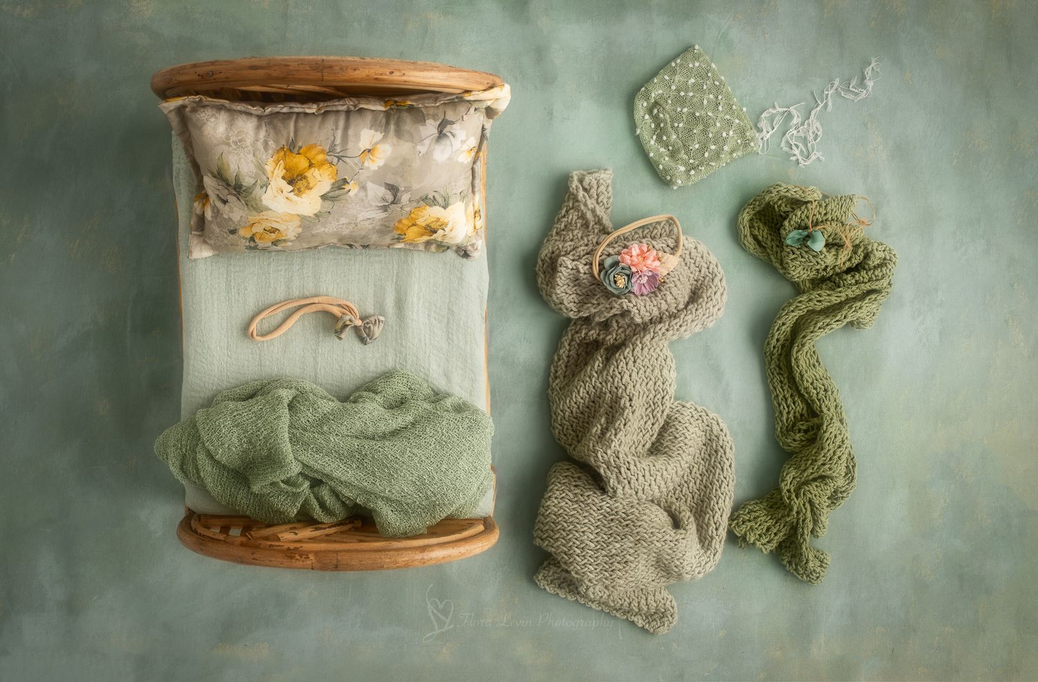 Flat lay of a miniature wooden bed with green knit wrap, floral pillow, and crochet newborn props on teal background.