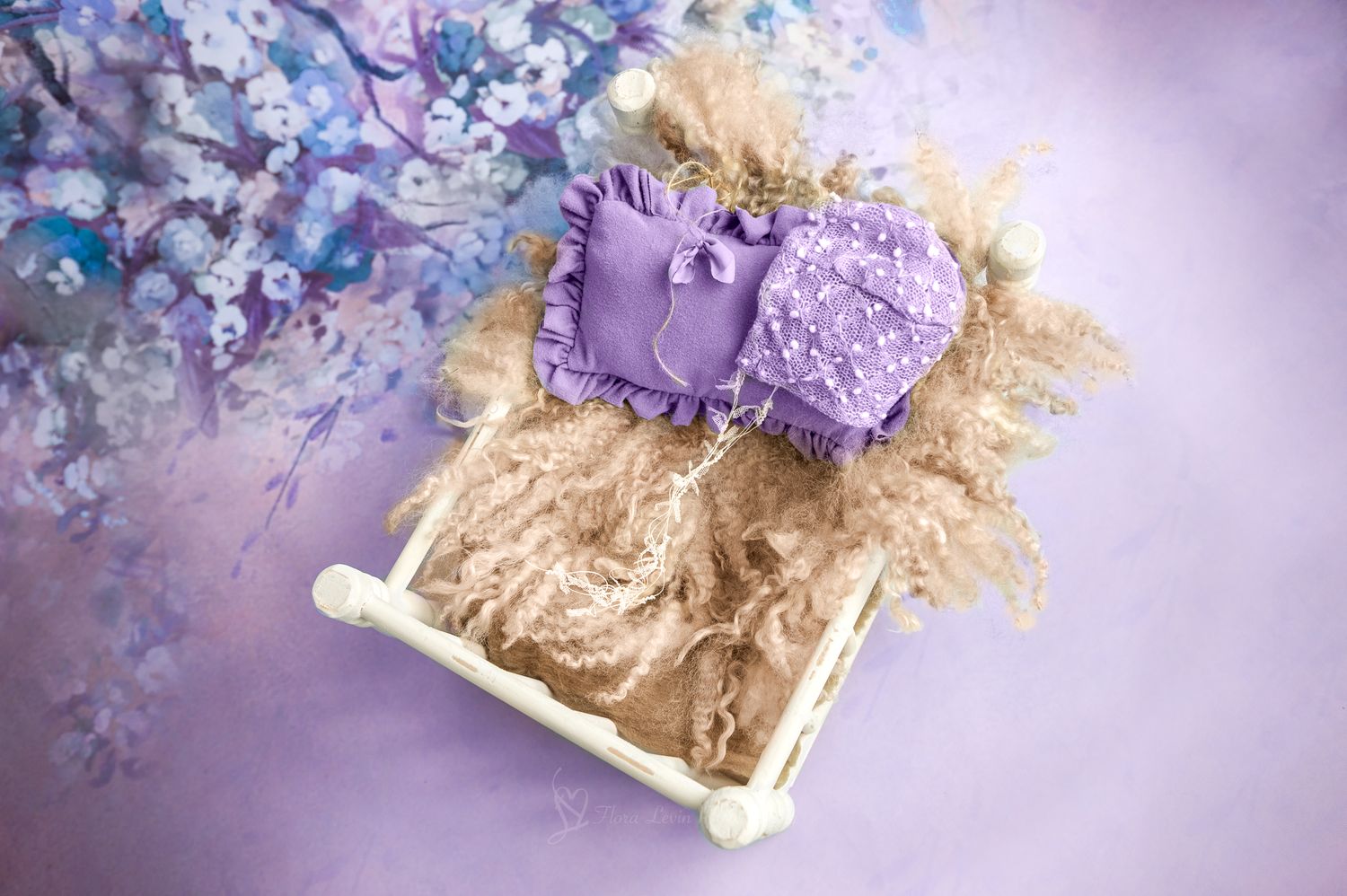 Purple lace newborn headband and pillow set displayed in a white mini crib with fluffy tan filler on a lavender with blue floral background.