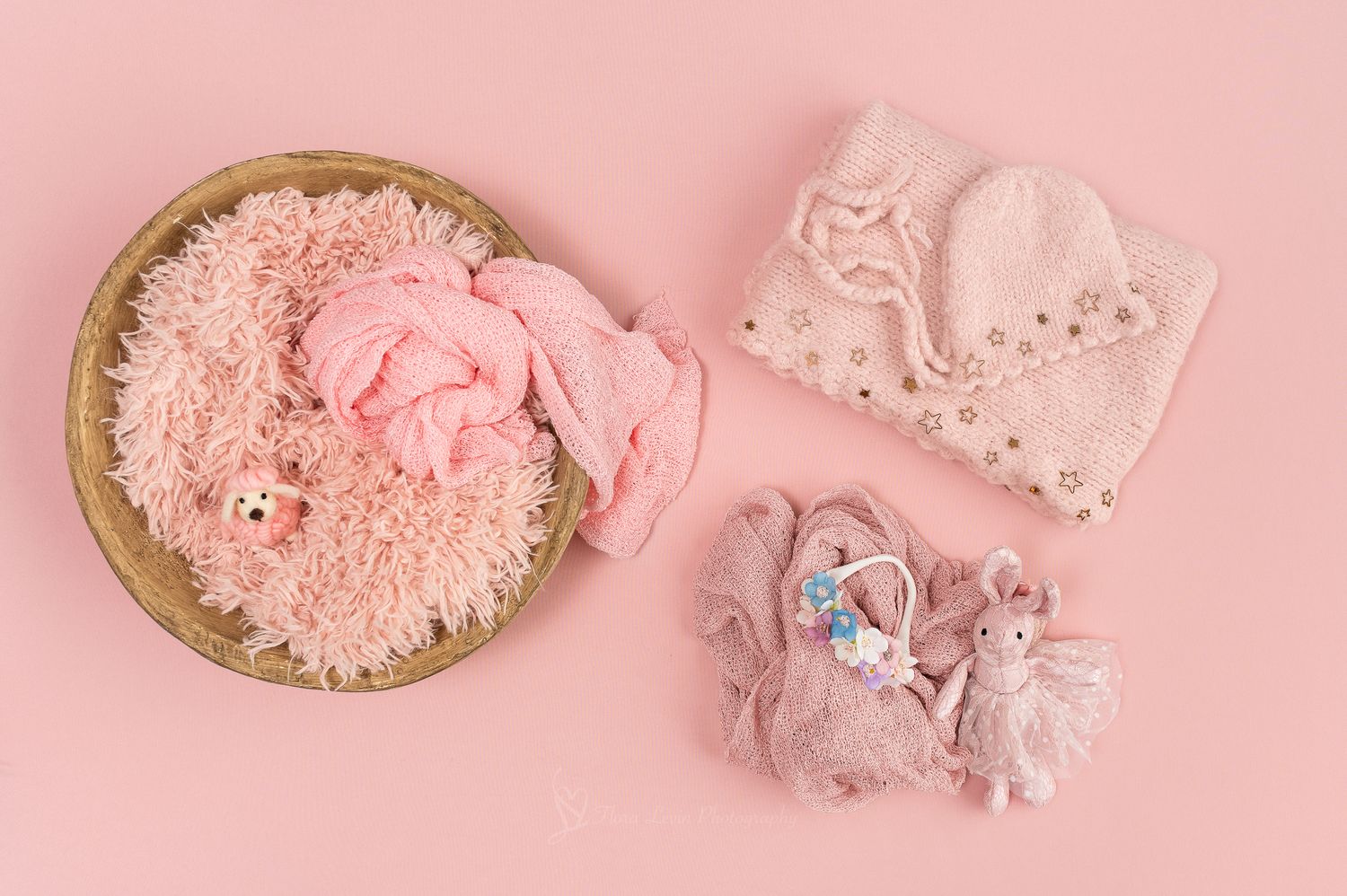 Pink newborn photography props including a fluffy bowl, knit hat and wrap, and bunny toys on a pink background.
