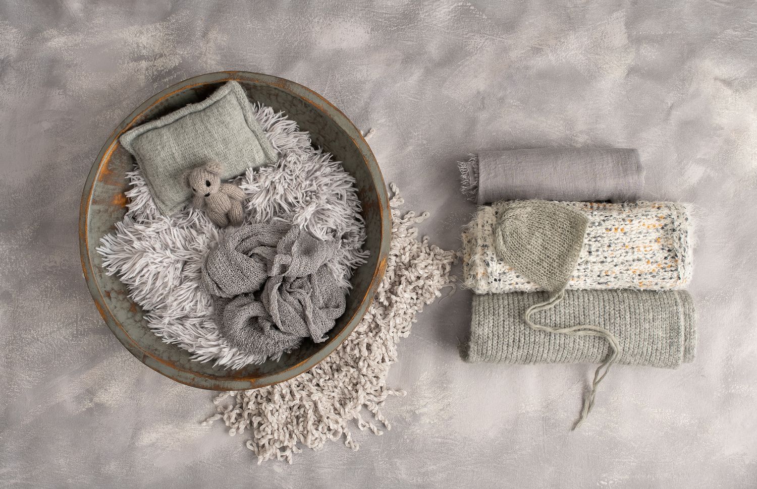 Neutral gray newborn photography props including knit blankets, hat, and bowl with fluffy gray filler on gray backdrop.