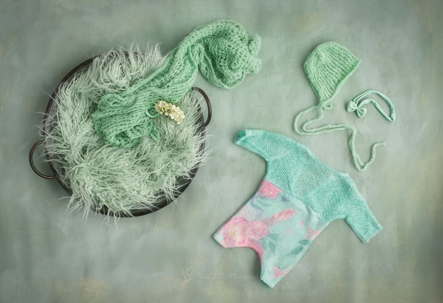 Mint green newborn photography props including knit bonnet, wrap, fluffy basket filler, and floral outfit on green background.